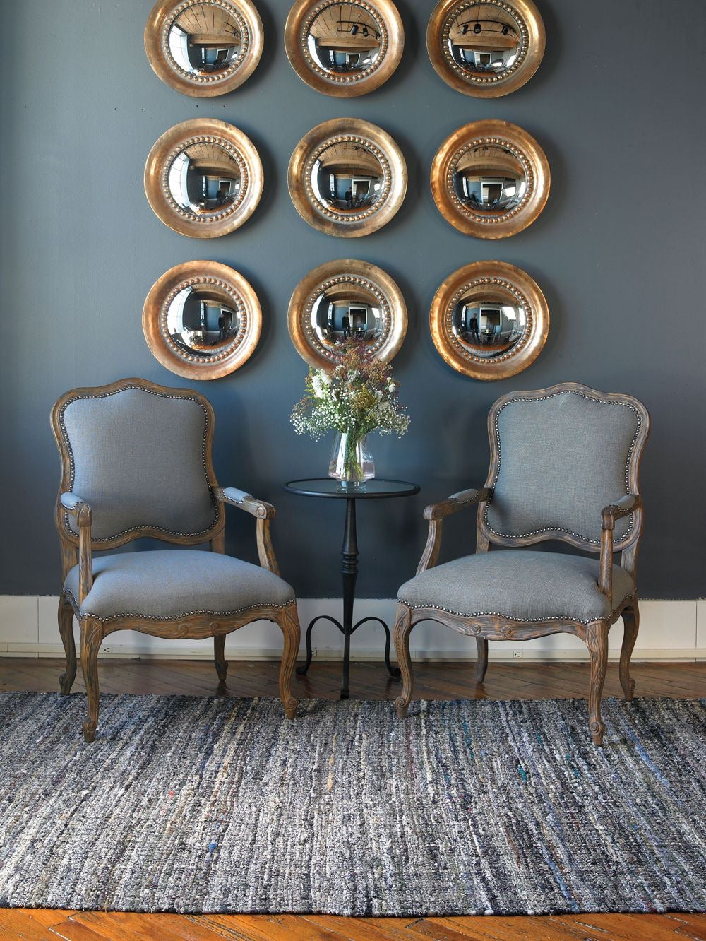 A pair of rustic French Country style armchairs with natural wood frames and grey upholstered seats, flanking a small metal table with flowers