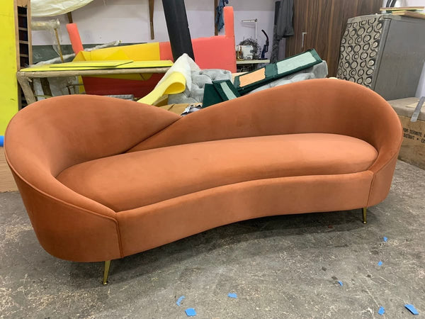  A luxurious terracotta velvet sofa with a unique comma shape and slender gold legs, set in a workshop or production area.