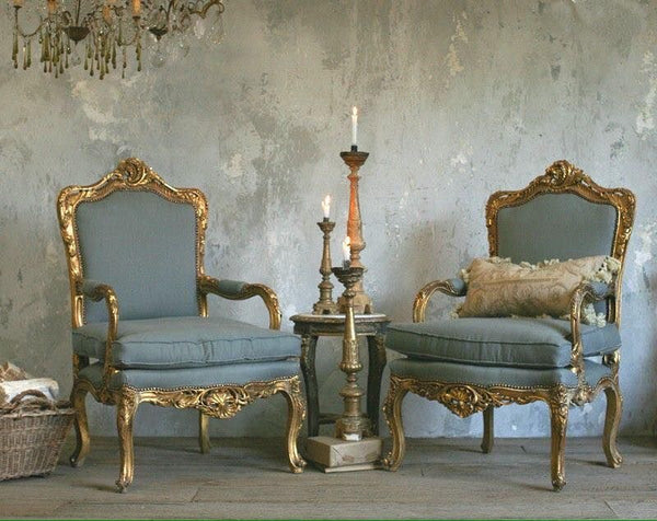 A pair of ornate antique gold French armchairs with blue upholstery, flanking a small side table with lit candlesticks, set against a textured gray wall, under a chandelier.