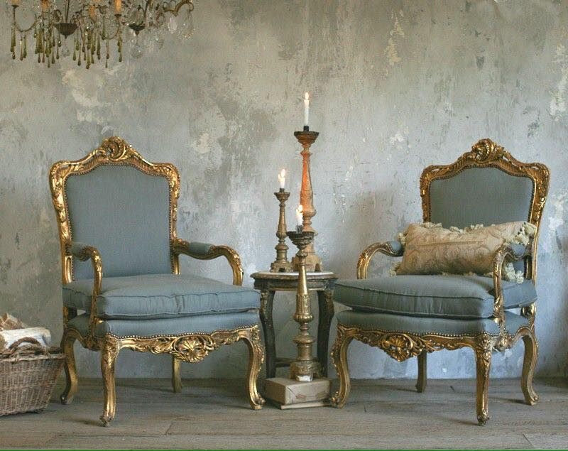 A pair of ornate antique gold French armchairs with blue upholstery, flanking a small side table with lit candlesticks, set against a textured gray wall, under a chandelier.