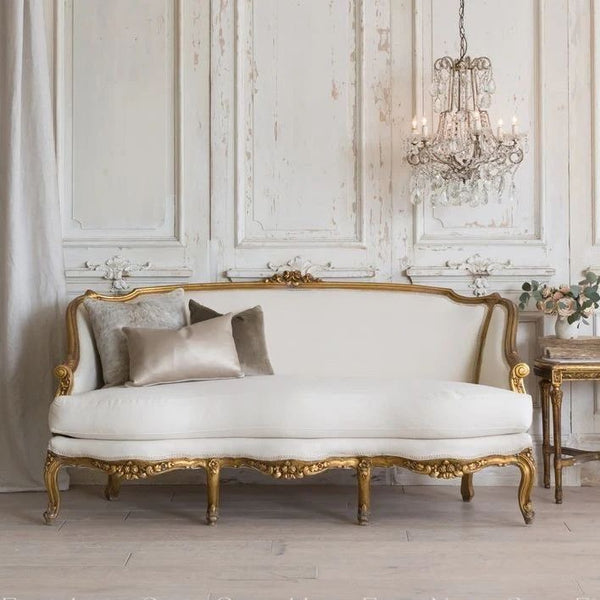 A Louis XV style sofa with an ornate gold carved frame, upholstered in cream fabric, and accented with two decorative throw pillows.