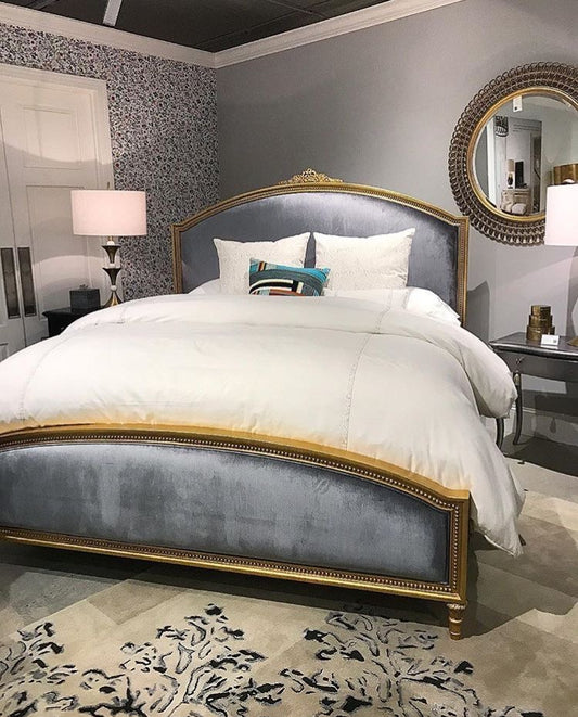A luxurious bed with a wide, arched headboard and footboard upholstered in grey velvet, set within a gilded gold frame, dressed with white bedding and accent pillows.