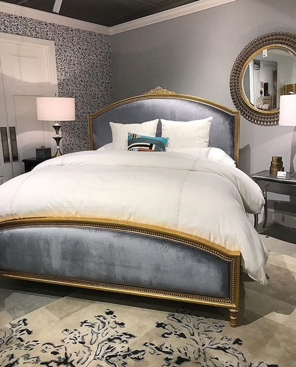 A luxurious bed with a wide, arched headboard and footboard upholstered in grey velvet, set within a gilded gold frame, dressed with white bedding and accent pillows.