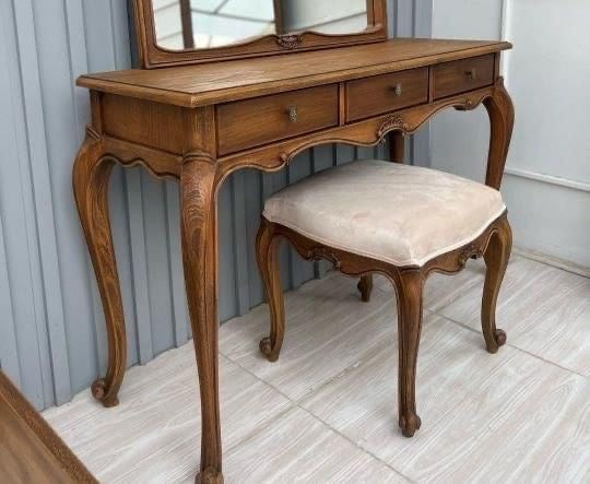 A wooden French-style dressing table with three drawers, elegant cabriole legs, a decorative mirror, and a matching upholstered vanity stool.
