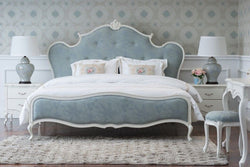 A French country style bed with a blue upholstered, tufted headboard and footboard, white frame, floral patterned bedding