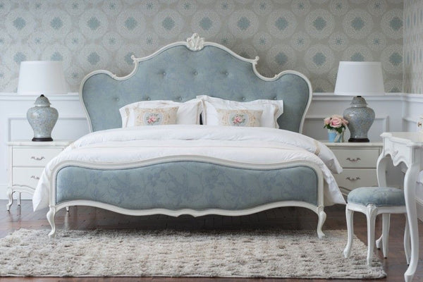 A French country style bed with a blue upholstered, tufted headboard and footboard, white frame, floral patterned bedding