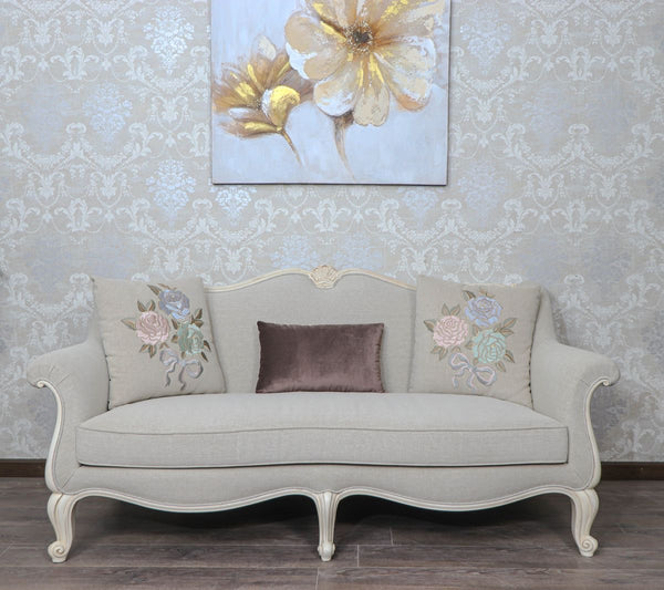 Provencal style 3-seater sofa in light grey with carved white wood frame and floral embroidered pillows.
