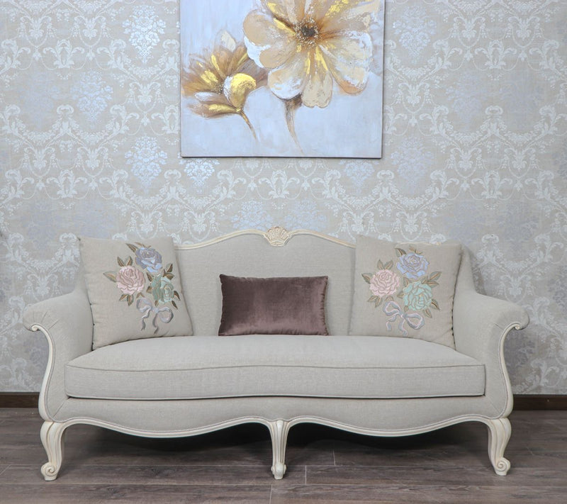 Provencal style 3-seater sofa in light grey with carved white wood frame and floral embroidered pillows.
